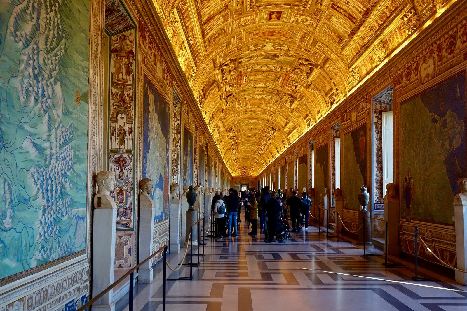 The Gallery of Maps in the Vatican Museums: a long corridor under a gilded, painted vault, lined with painted maps of Italy and marble busts, with visitors walking down the middle