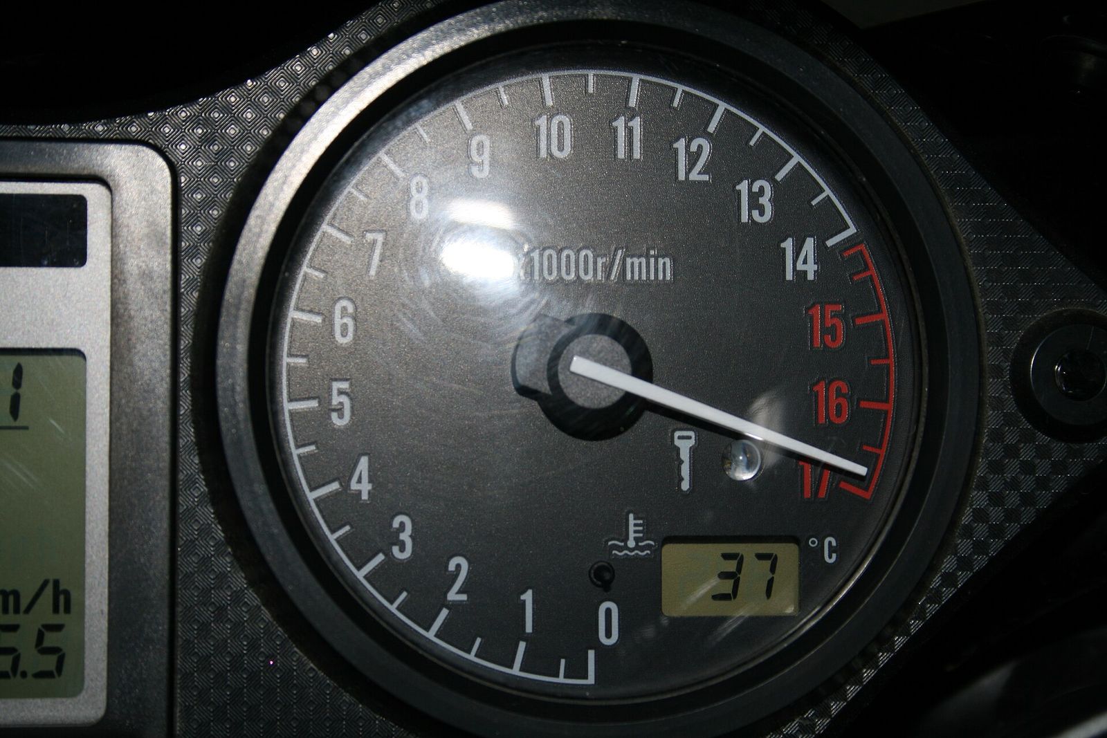 A motorcycle rev counter on a dark dial numbered 0 to 17, with its white needle pushed past the red band at the top of the scale