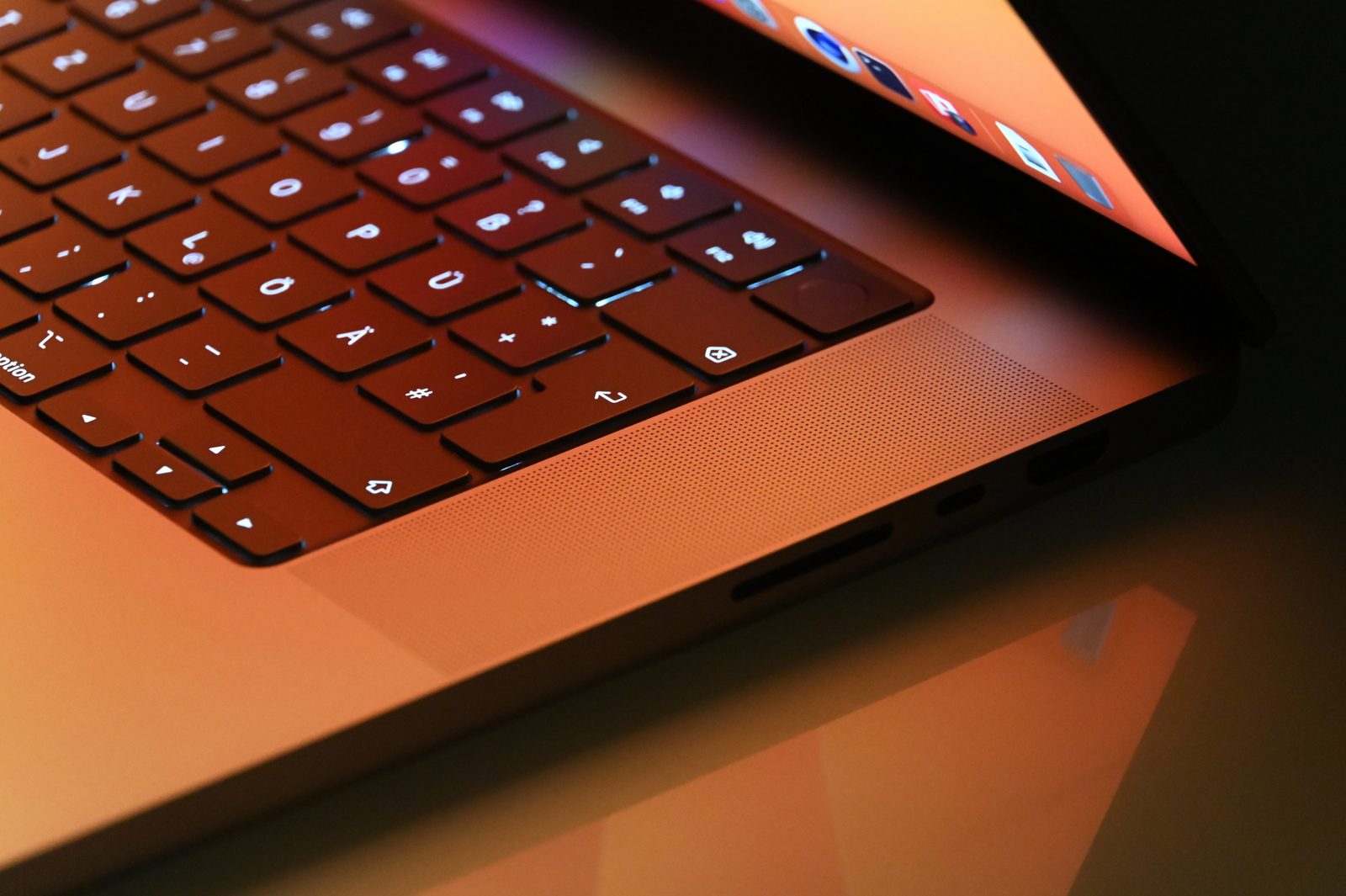 Close-up of a MacBook Pro's backlit keyboard and speaker grille, lit orange by its open screen and reflected in a glossy desk