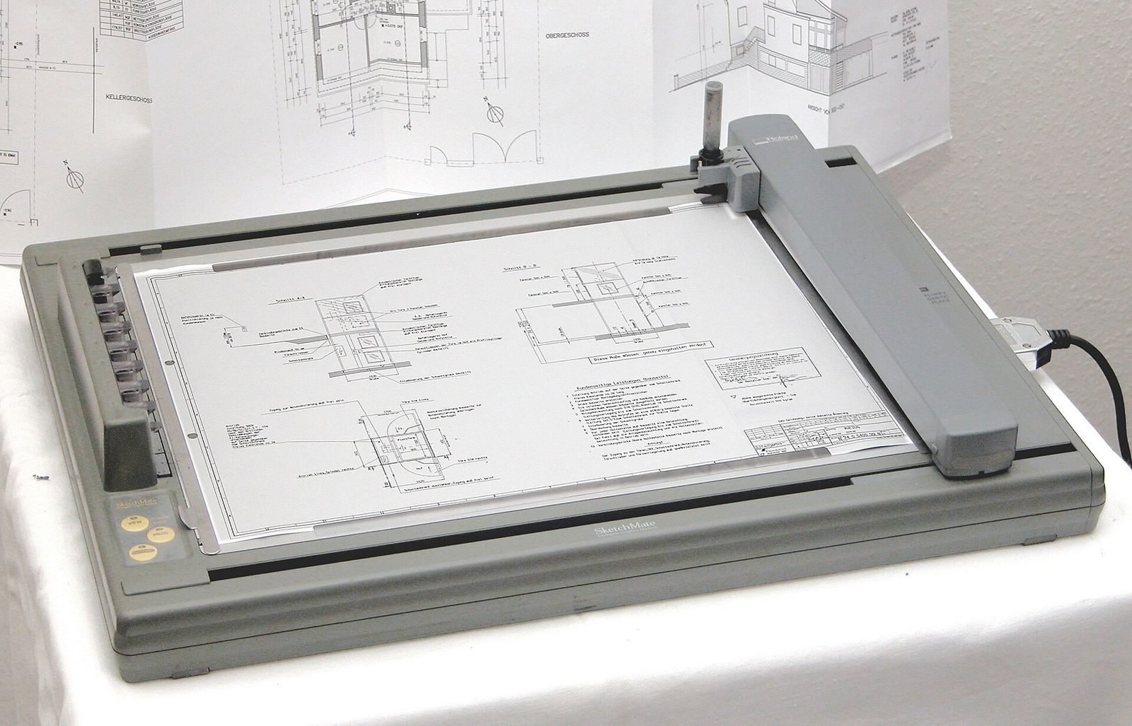 A grey flatbed pen plotter on a white table, with a printed building plan clamped to its bed and more plans hanging behind it
