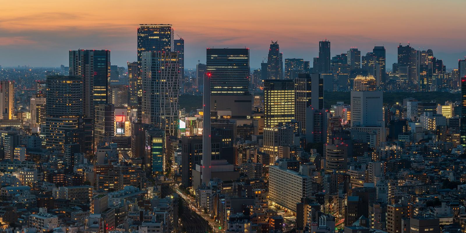 Tokyo at dusk from a high floor in Ebisu: the lit towers of Shibuya in the foreground and the skyscrapers of Shinjuku along the horizon under an orange sky