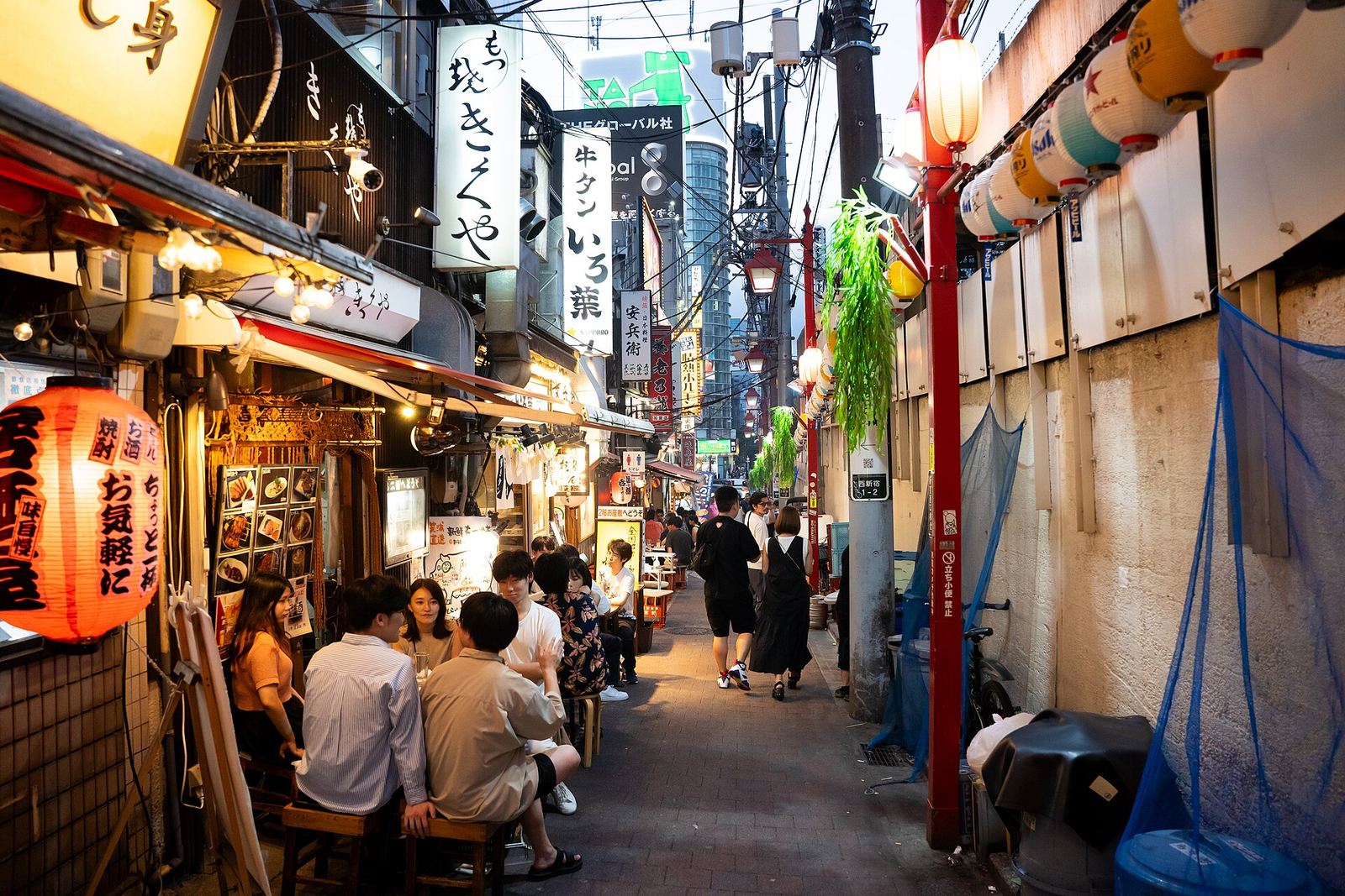 A narrow lane in Omoide Yokocho lined with tiny restaurants, red lanterns and hanging signs, with people eating at an open-fronted counter