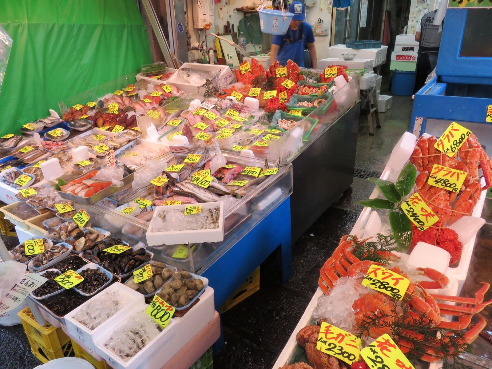A seafood stall in the Tsukiji Outer Market with trays of shellfish, fish and red crabs under handwritten yellow price tags