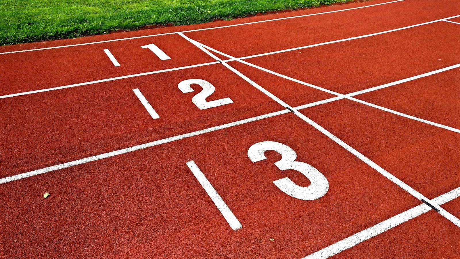 Lane numbers 1, 2 and 3 painted in white at the start line of a red running track, with a strip of grass beyond the first lane