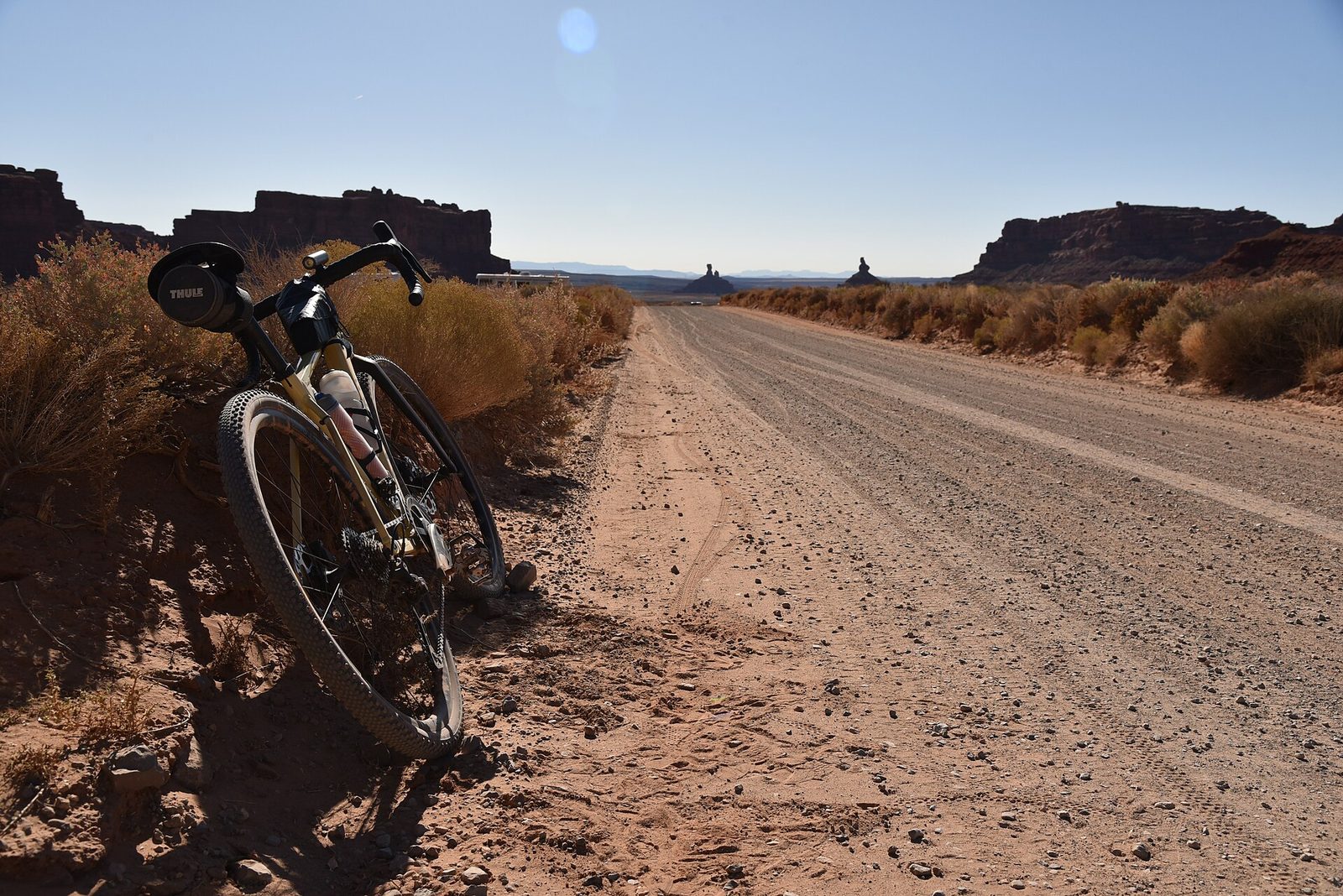 A gravel bike leaning against desert scrub at the side of an empty dirt road, with sandstone buttes on the horizon